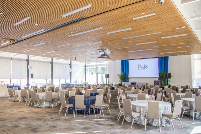 Photo of Penn Pavilion Room showing round tables, chairs, stage, windows, and projection screen.